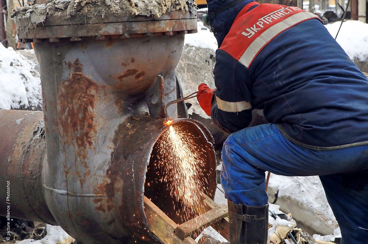 A worker in a padded winter jacket crouches beside a large industrial pipe or valve assembly, using a cutting torch that sends bright orange sparks flying near an open pipe opening. The scene is set outdoors in snowy conditions, with rusted metal piping visible in the background.