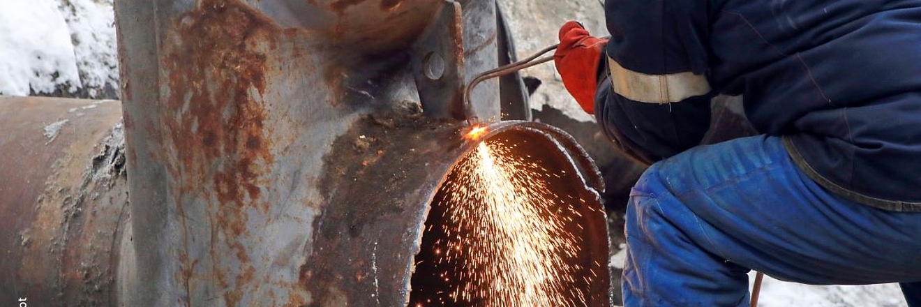 A worker in a padded winter jacket crouches beside a large industrial pipe or valve assembly, using a cutting torch that sends bright orange sparks flying near an open pipe opening. The scene is set outdoors in snowy conditions, with rusted metal piping visible in the background.