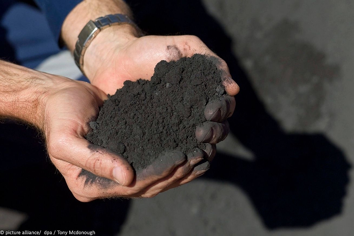 "Two hands holding a pile of dark, fine-grained mineral powder or ore concentrate, with fingers stained black from the material."