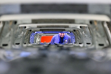 "View through a series of stacked, unpainted steel car body shells on a production line, creating a tunnel-like perspective. In the centre, a smiling worker in a blue uniform and cap is visible, inspecting the vehicles. Photo: picture alliance / dpa / Julian Stratenschulte."