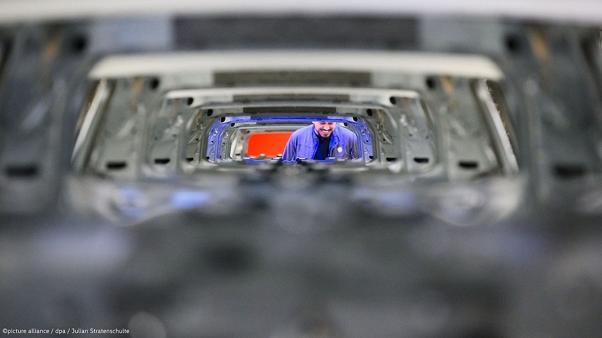 "View through a series of stacked, unpainted steel car body shells on a production line, creating a tunnel-like perspective. In the centre, a smiling worker in a blue uniform and cap is visible, inspecting the vehicles. Photo: picture alliance / dpa / Julian Stratenschulte."