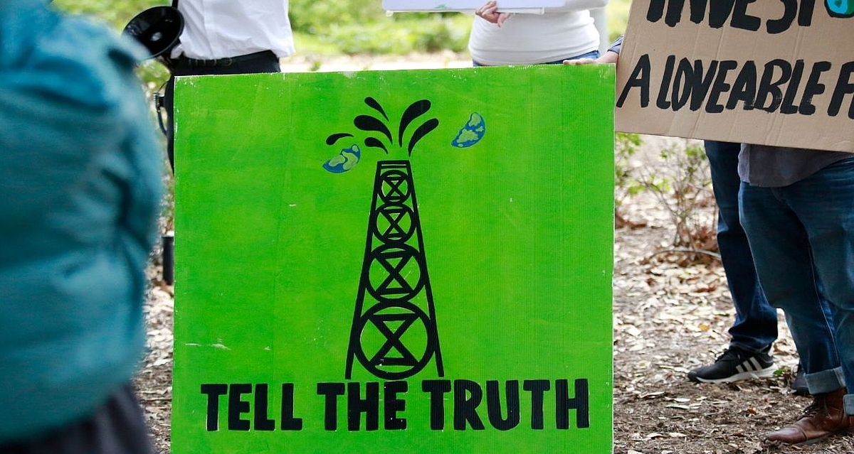 "Climate activist holding a green protest sign reading 'Tell the Truth', featuring an oil derrick with the Extinction Rebellion symbol, at an outdoor demonstration."