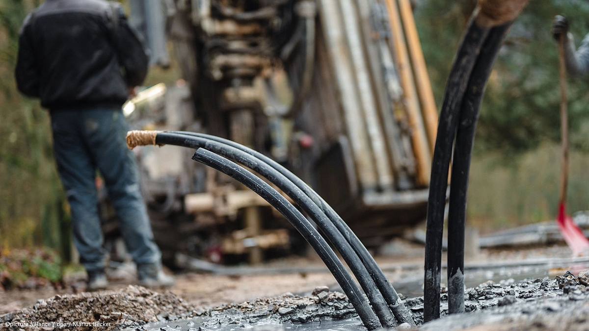 "Geothermal drilling equipment with curved black hoses in the foreground and a worker operating a drilling rig in the background"