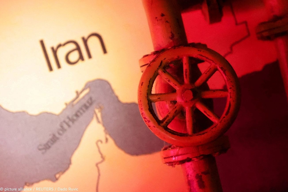 Close-up of an aged, rust-coloured pipeline valve bathed in red light, positioned in front of a map. The map shows the region around Iran with the label "Iran" clearly visible, and the Strait of Hormuz marked in the lower left. The red lighting creates a tense, warning atmosphere. Photo credit: picture alliance / REUTERS / Dado Ruvic.