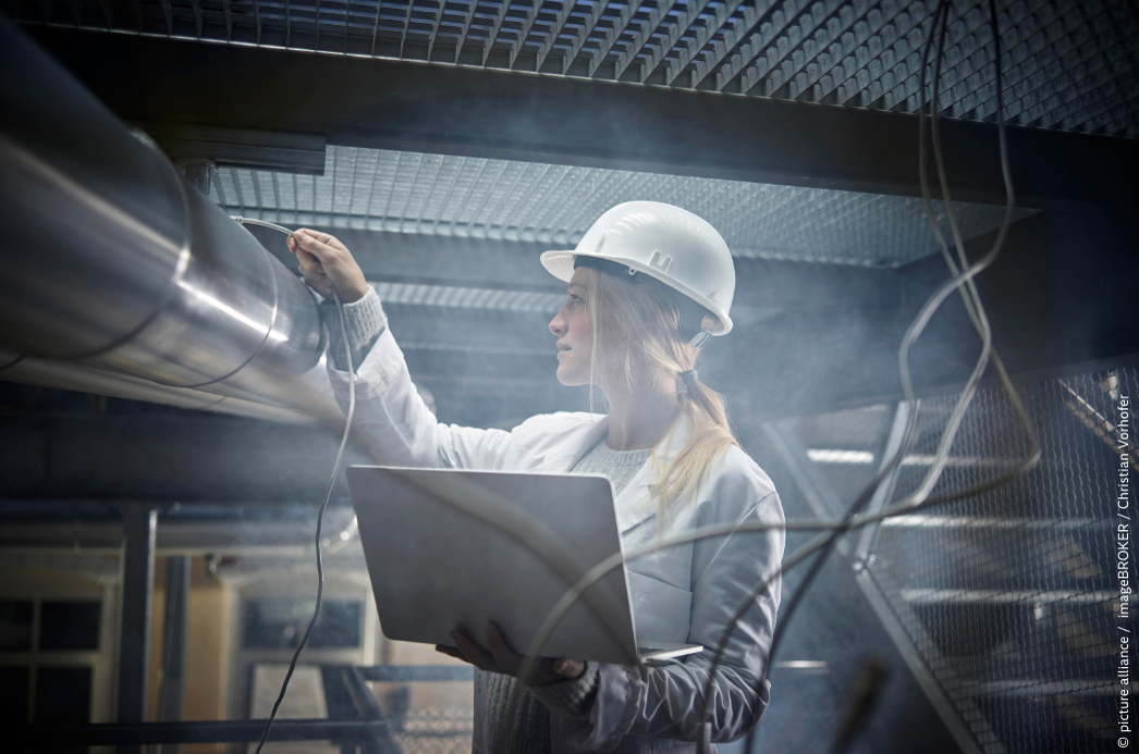  A young woman with long blonde hair, wearing a white hard hat and a white lab coat, holds an open laptop in one hand while reaching up to inspect large silver industrial pipes or ducts with the other. She is working in an industrial facility with visible cables, metal grating, and a hazy atmosphere suggesting steam or dust. Her focused expression suggests she is conducting a technical inspection or measurement. Photo credit: picture alliance / imageBROKER / Christian Vorhofer.