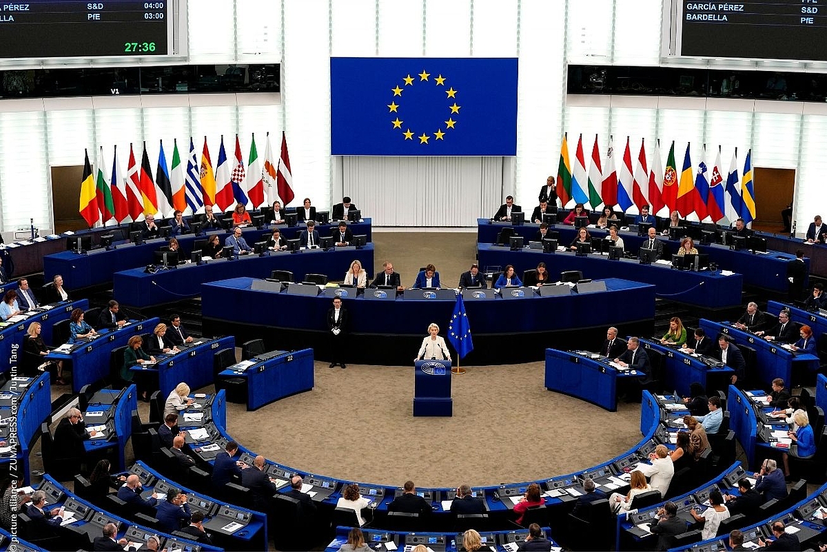 European Commission President Ursula von der Leyen delivers the State of the European Union address from the podium of the European Parliament's hemicycle in Strasbourg, surrounded by seated MEPs, EU member state flags and a large EU flag displayed on the screen behind her.