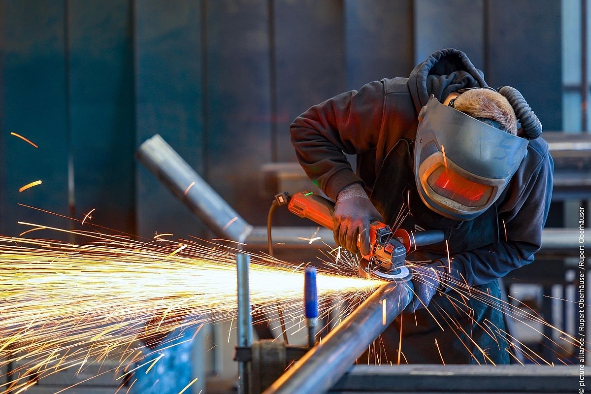 The image shows a worker grinding metal with an angle grinder, sparks flying dramatically — powerful visual for May Day and industrial policy themes