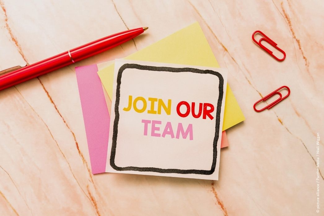 The picture shows a red ballpoint pen and two red paper clips wrapped around a small stack of Post-it notes, one of which reads ‘Join our team’