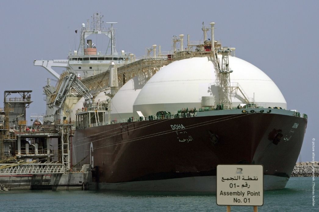 An LNG tanker named "Doha" docked at a liquefaction terminal in the Gulf region. The vessel's large spherical storage tanks are visible alongside port infrastructure and an "Assembly Point No. 01" safety sign in Arabic and English.