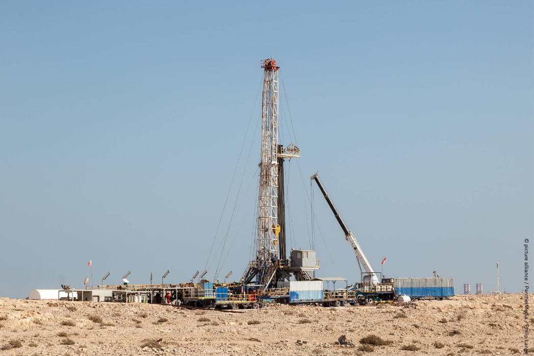 Oil drilling rig in a barren desert landscape under a cloudless sky, surrounded by industrial equipment and a crane.